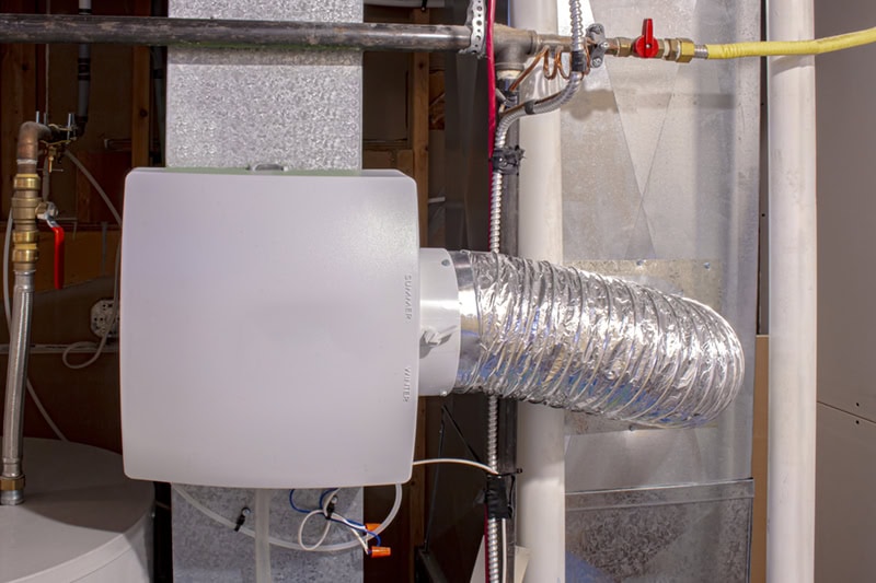 A white, rectangular humidifier unit attached to a sheet metal duct, with a thin flexible aluminum duct connected to its right side and a red wire running vertically past it.