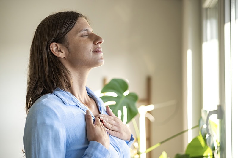A woman in a light blue collared shirt stands in profile facing right, with her eyes closed, a calm expression, and both hands resting on her chest.